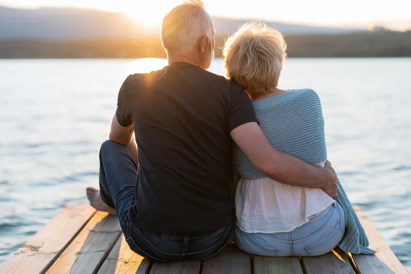 Elderly couple embracing while sitting on a wooden dock by a lake at sunset