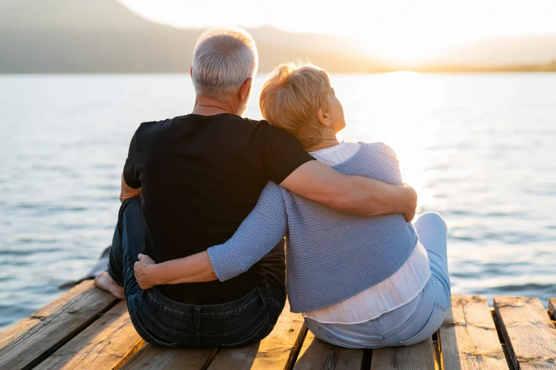 Senior couple embracing while sitting on a wooden dock by a lake at sunset