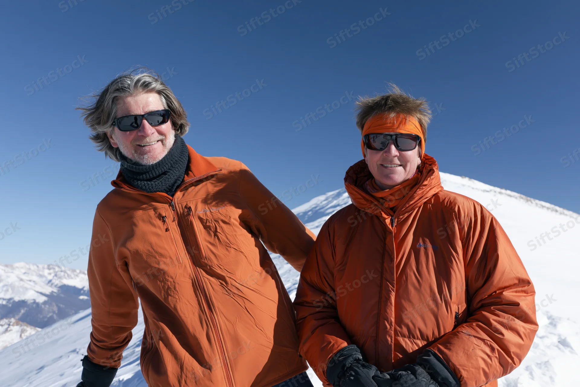 Two hikers in orange jackets smiling on a snowy mountain ridge under a clear blue sky