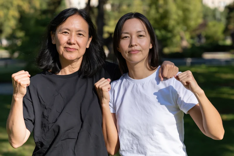 Two asian women posing together in a sunlit park, smiling with fists raised, wearing casual tshirts
