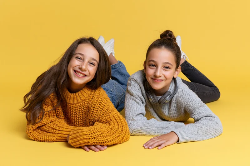 Two young girls lying on stomachs smiling at camera on yellow background