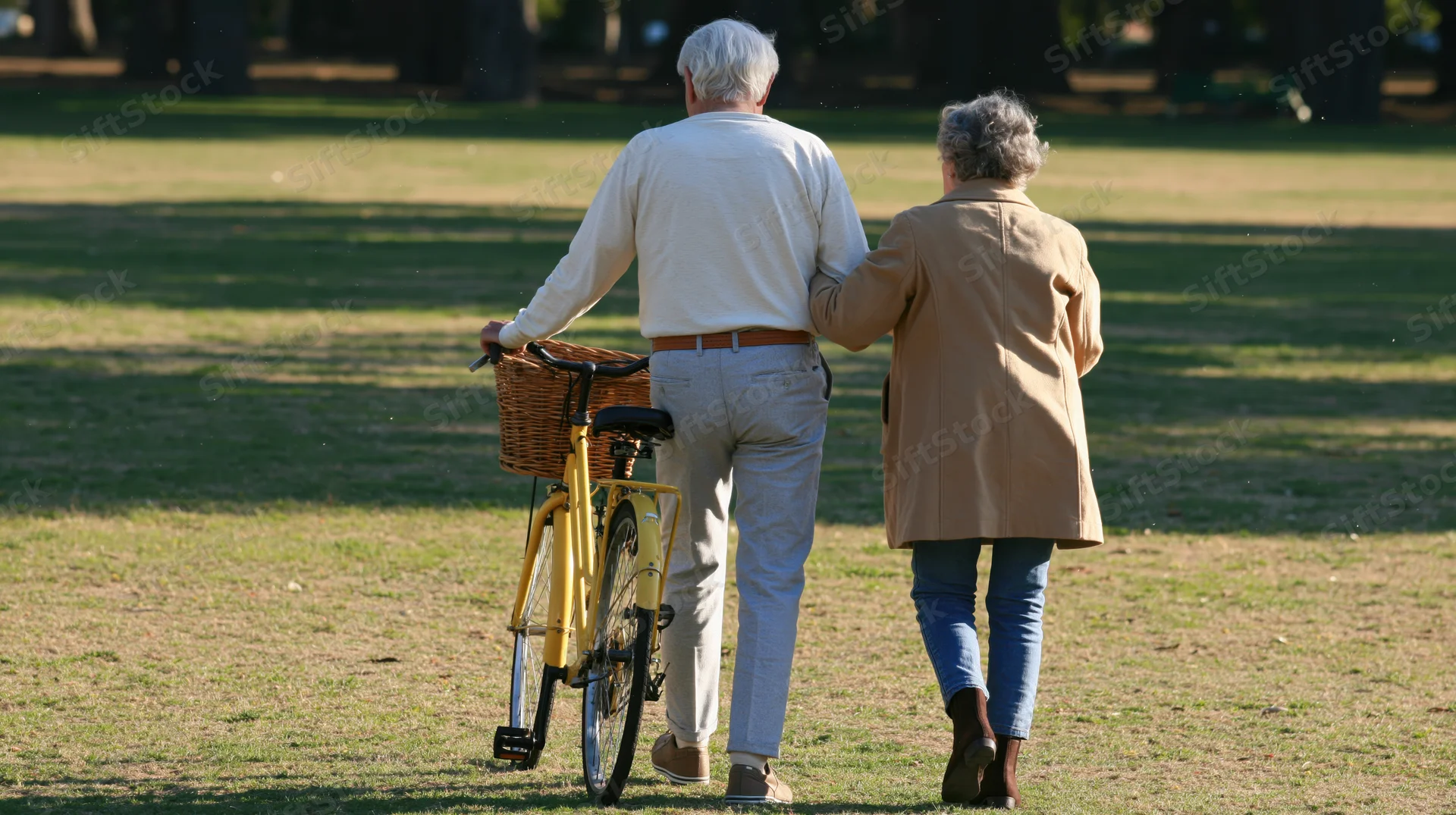 Elderly couple walking away across a grassy park with a bicycle, arm in arm