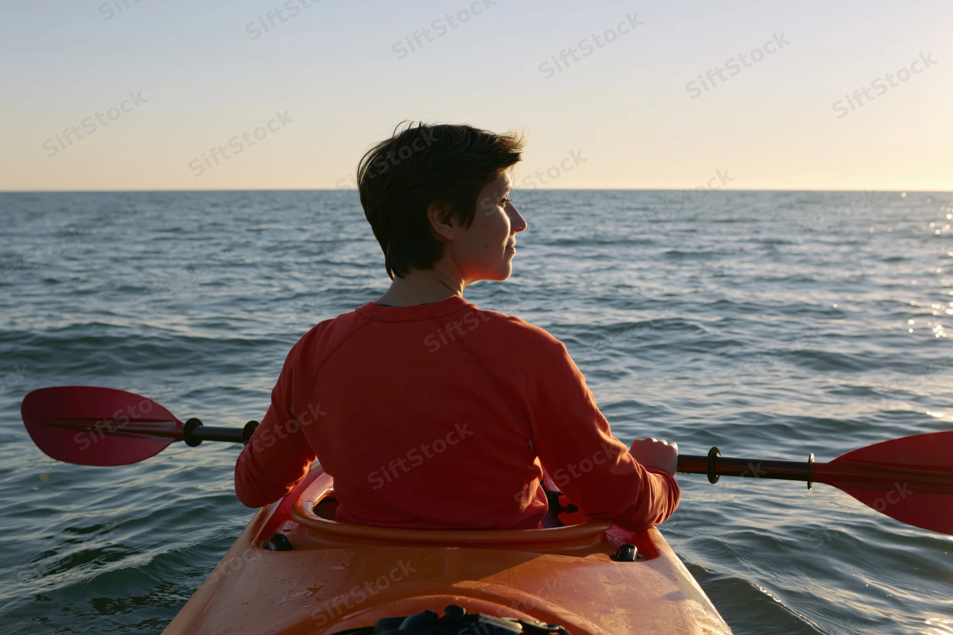 Woman kayaking on calm ocean at sunset, viewed from behind at water level