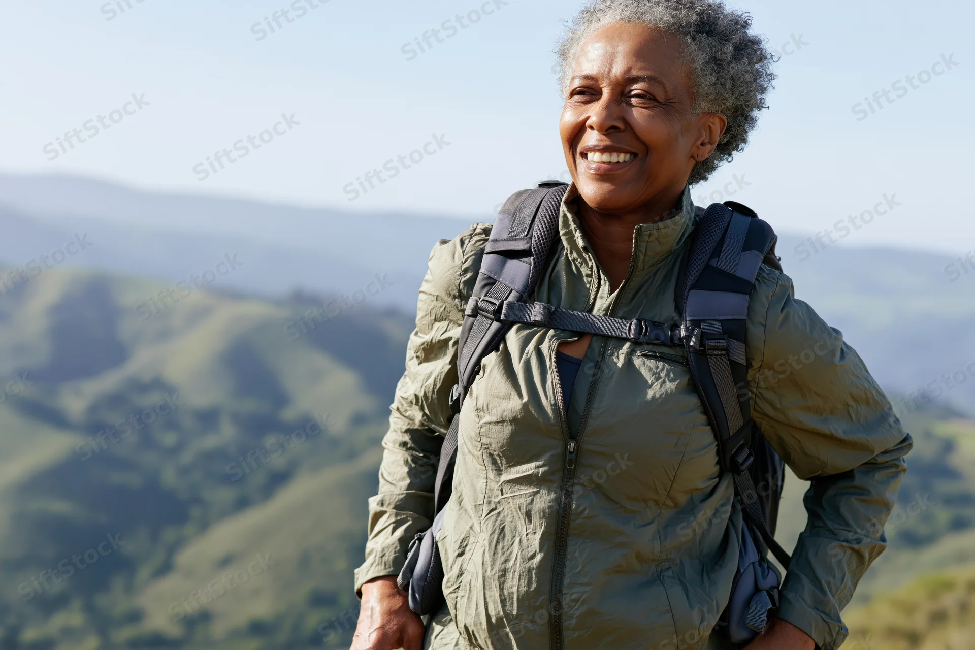 Senior black woman smiling with backpack on mountain trail overlooking green hills