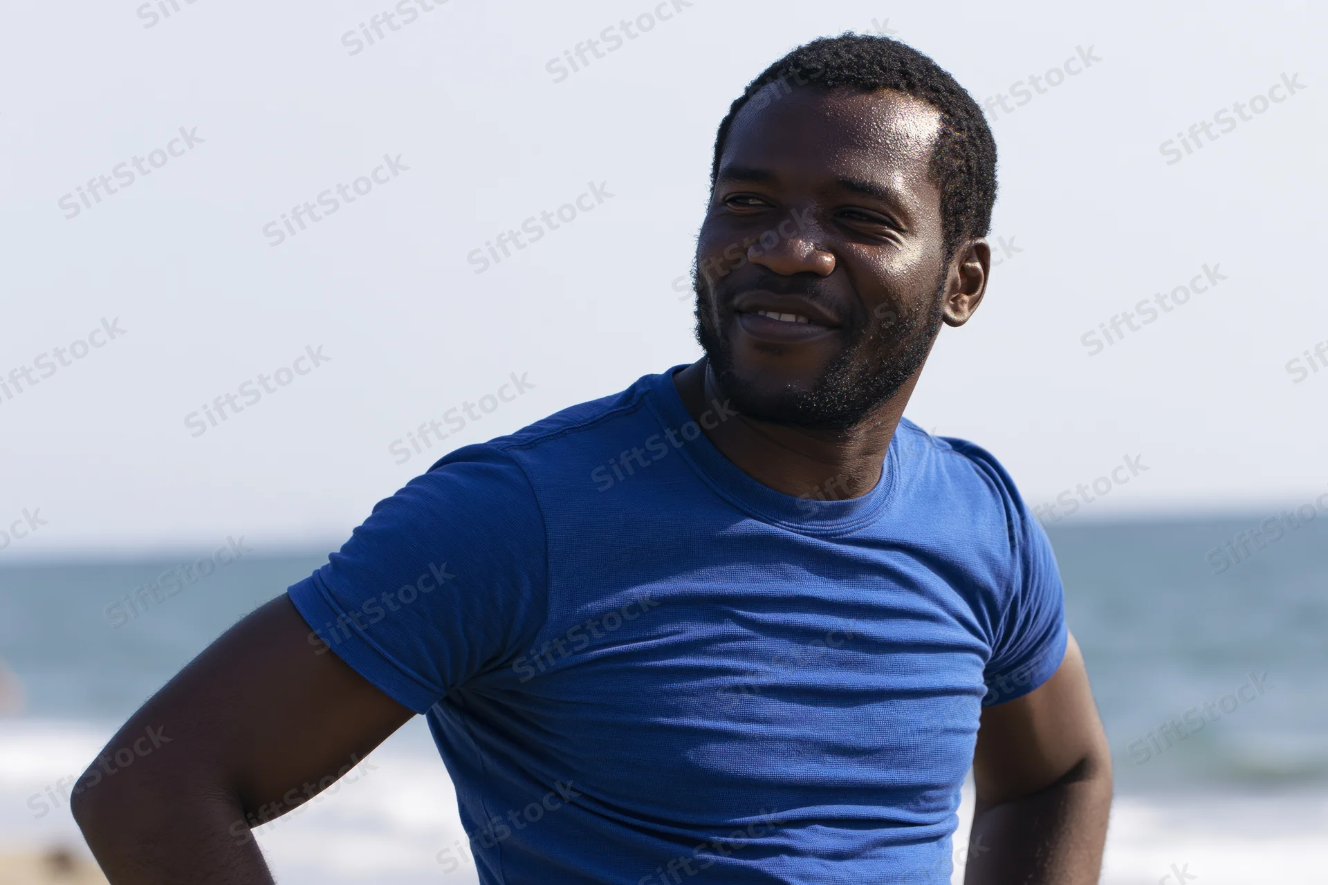 Smiling man in blue tshirt standing on sunny beach with ocean behind him
