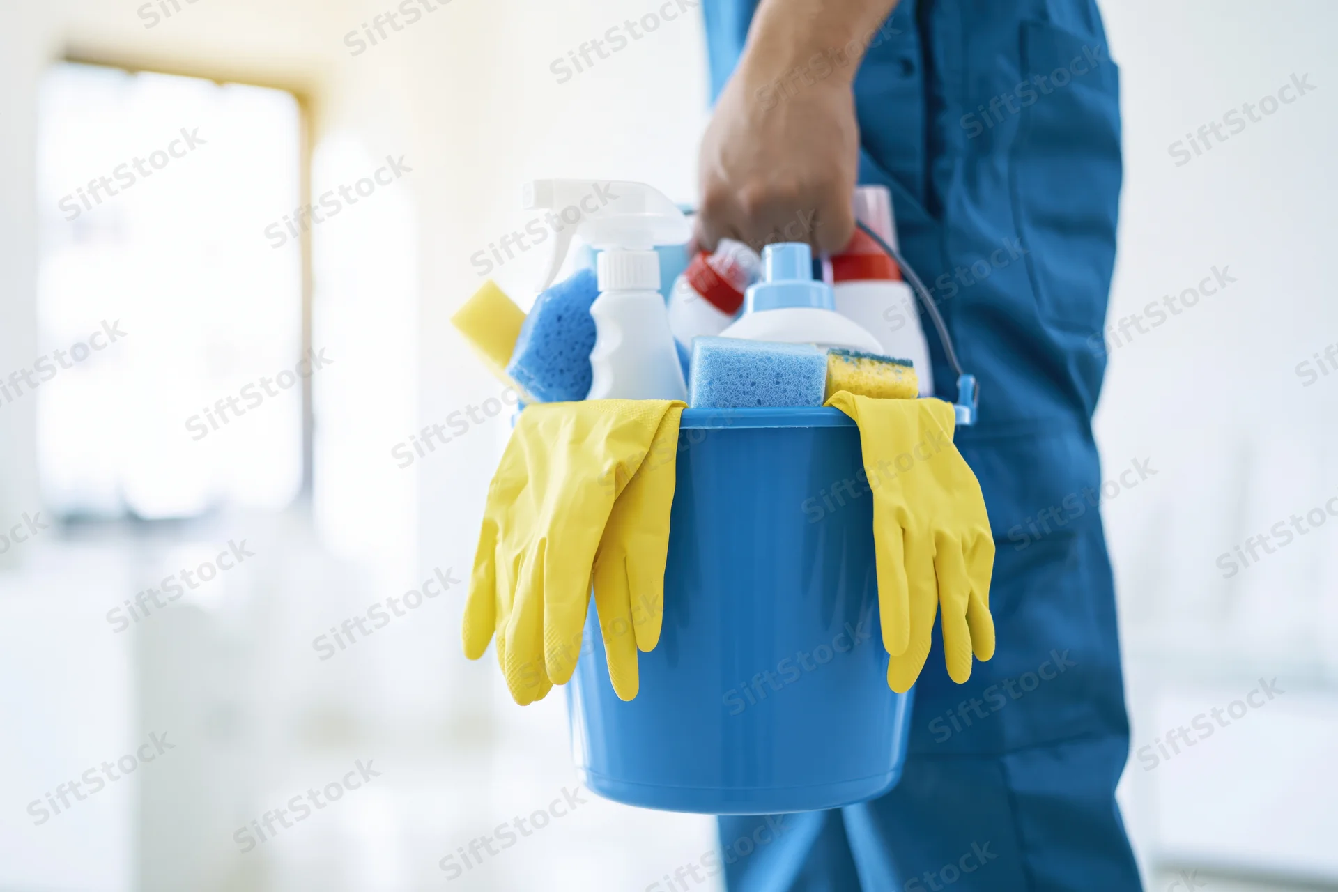 Man in blue uniform holding a blue bucket filled with cleaning supplies and yellow gloves indoors