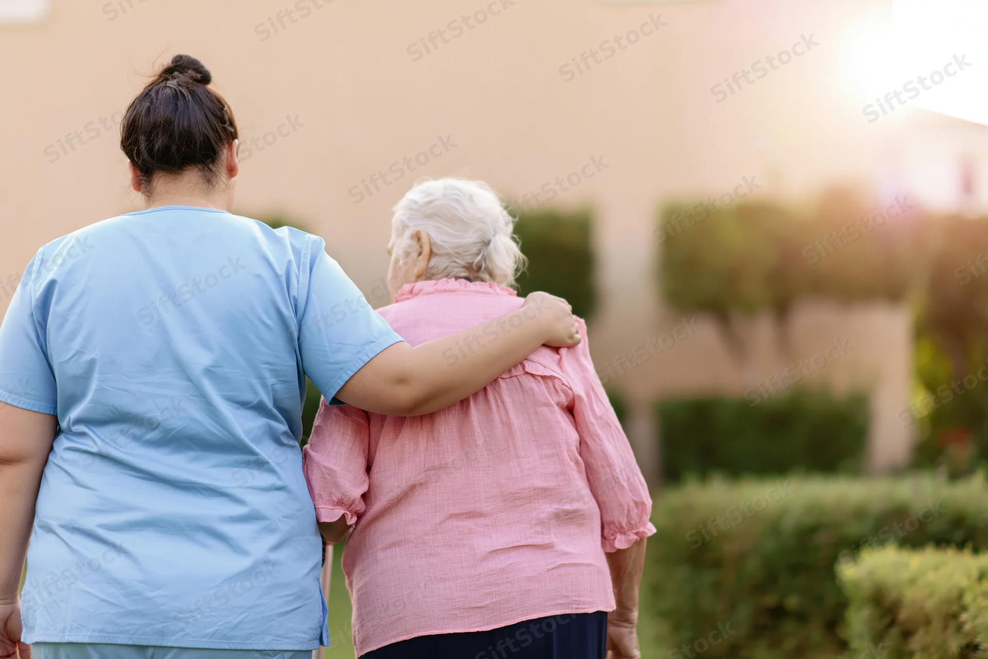 Caregiver walking arm-in-arm with senior woman in garden at sunset