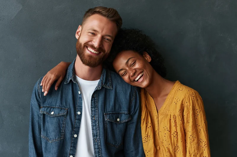 Young multiethnic couple smiling and laughing while leaning together against a dark grey textured wall