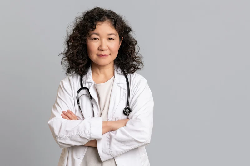 Asian woman doctor in white coat with stethoscope arms crossed standing in studio
