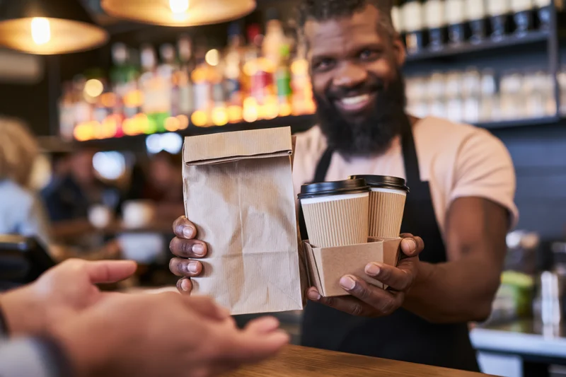 Bearded barista handing paper bag and coffee cups to customer at cafe counter