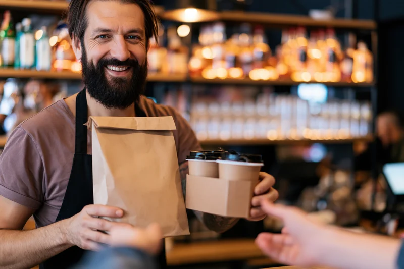 Bearded barista in black apron smiling while handing a brown paper bag and coffee cups to a customer at a cafe counter