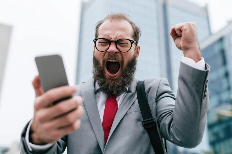 Bearded businessman in gray suit shouting in celebration while holding smartphone outdoors in city
