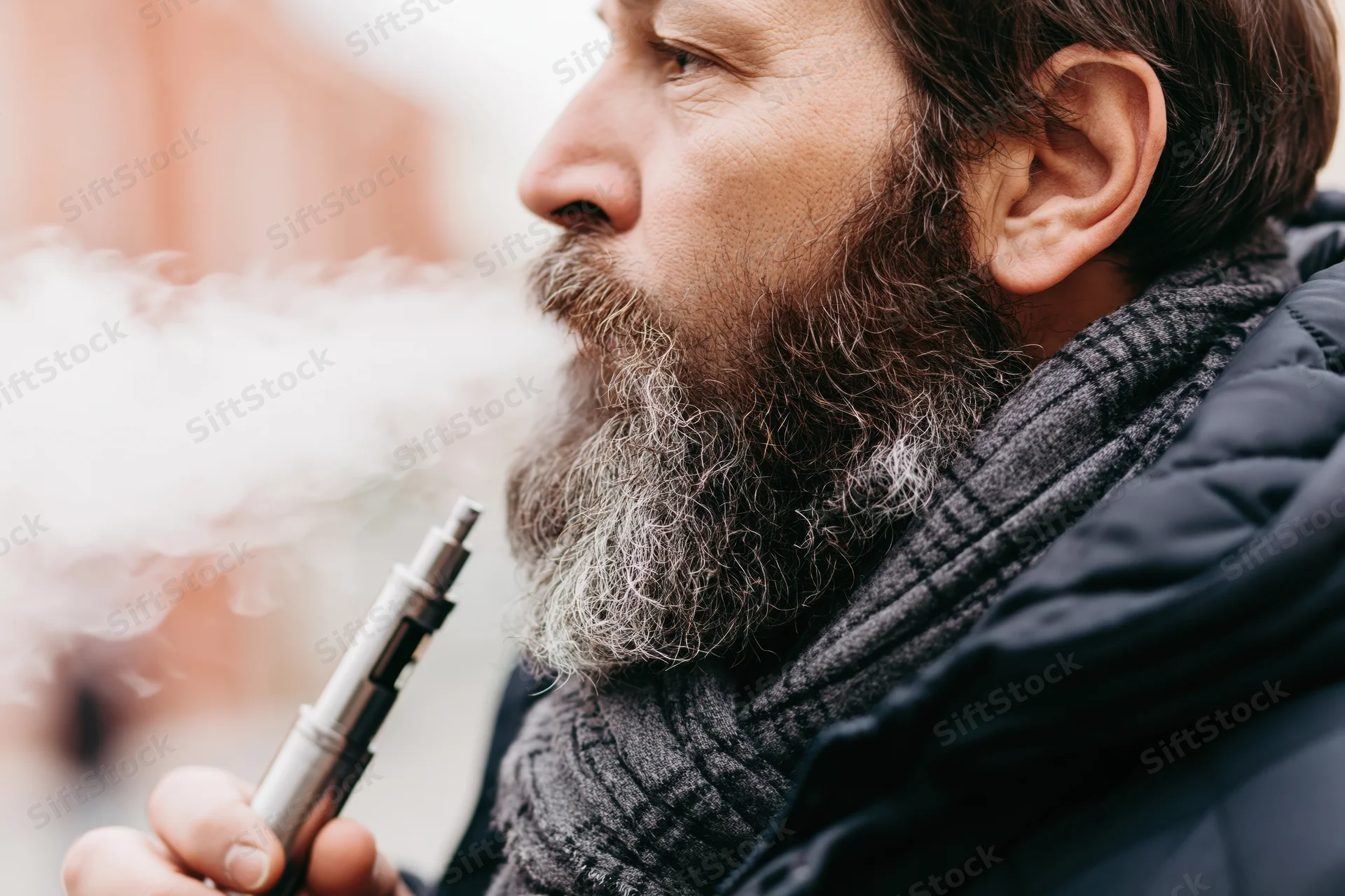 Bearded man exhaling vapor from metallic vape mod outdoors in winter clothing
