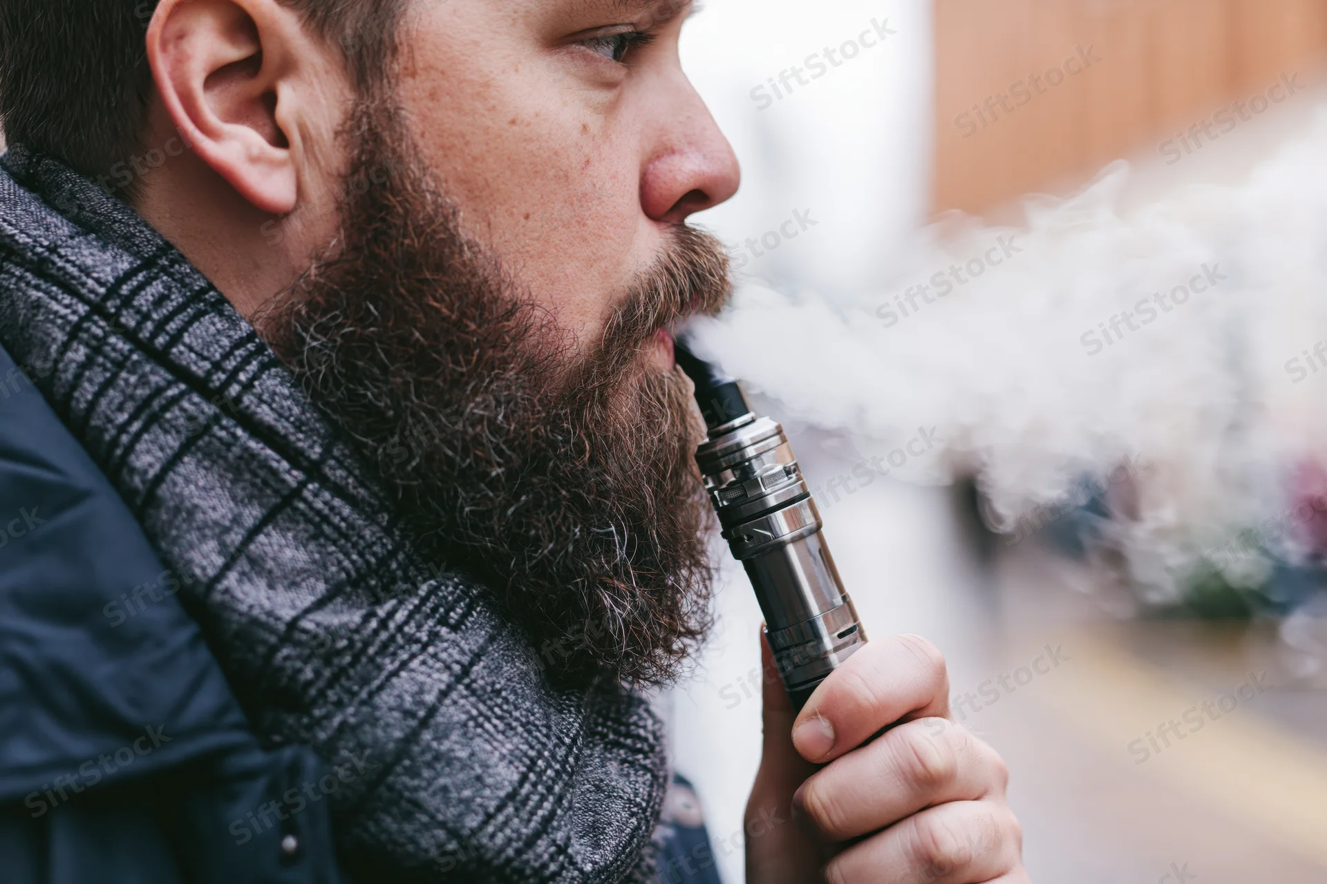 Bearded man exhaling vapor from metallic vape mod outdoors in winter coat and scarf