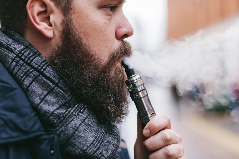 Bearded man exhaling vapor from metallic vape mod outdoors in winter coat and scarf