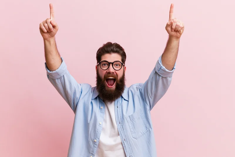 Bearded man celebrating with arms raised pointing upward against pink background