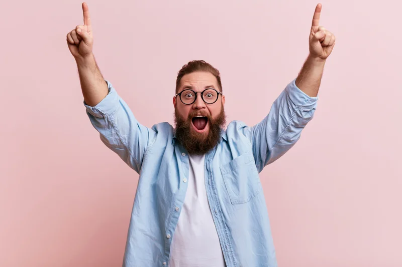 Bearded man celebrating with arms raised pointing upward against pink background