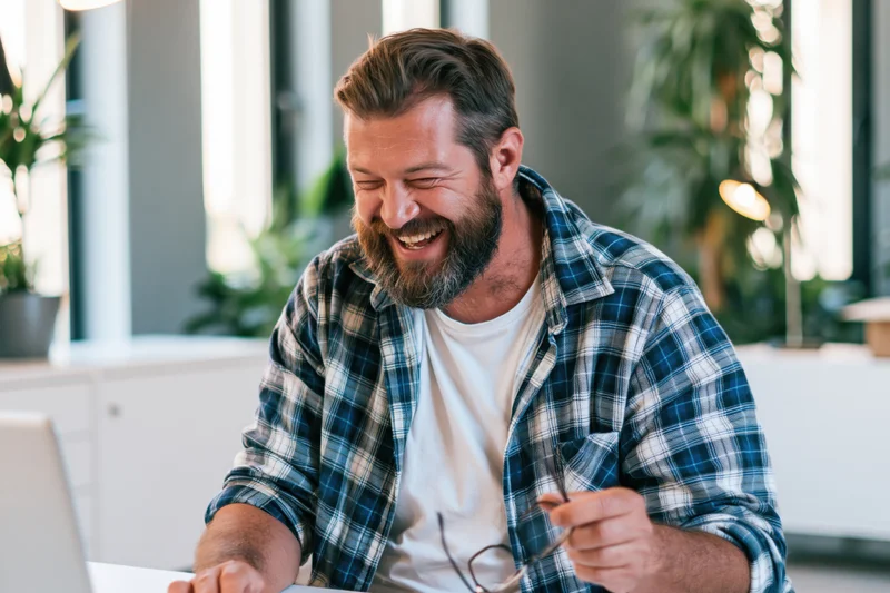 Bearded man laughing while using laptop at home office desk