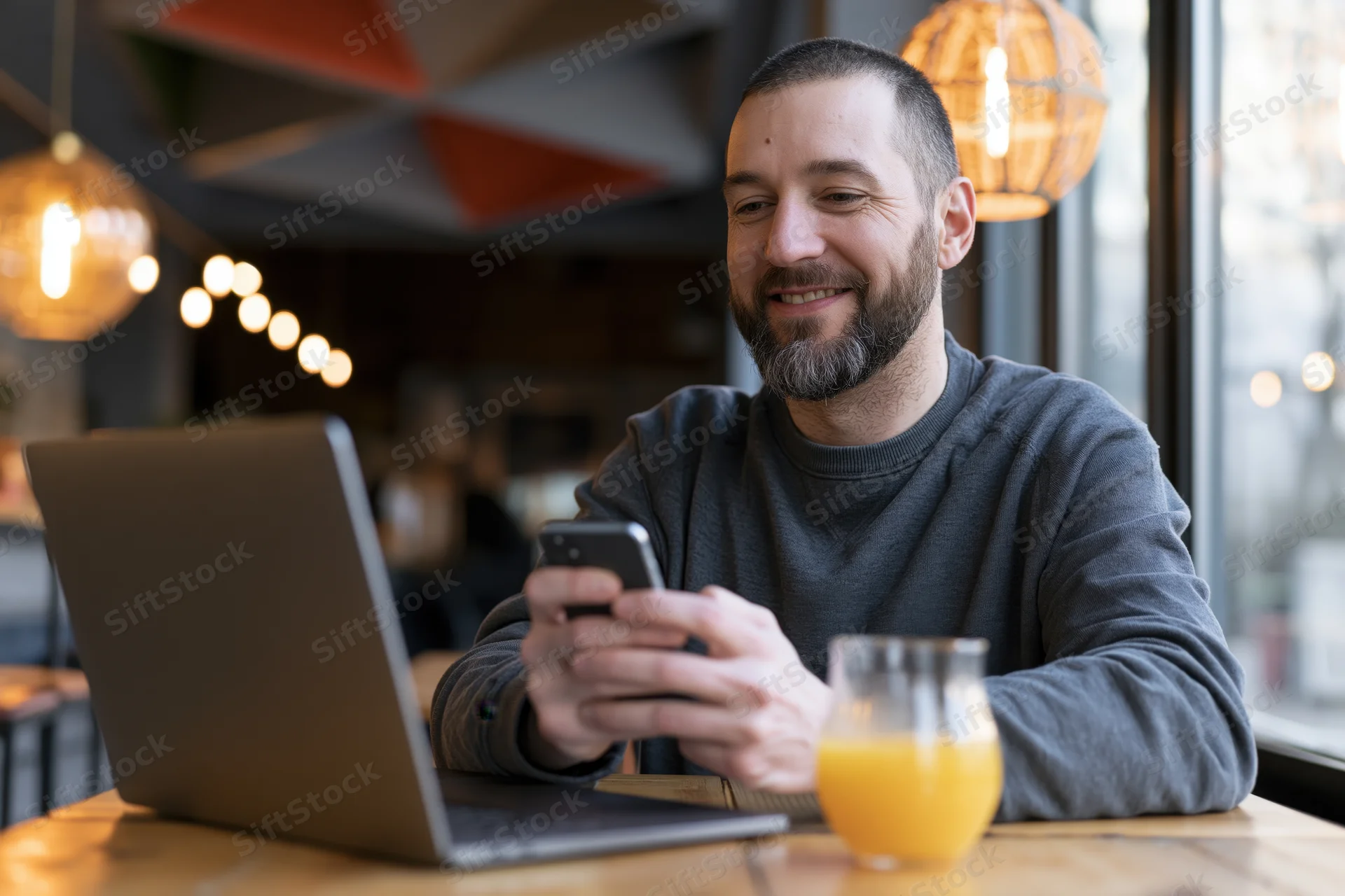 Bearded man smiling while using smartphone and laptop at wooden cafe table