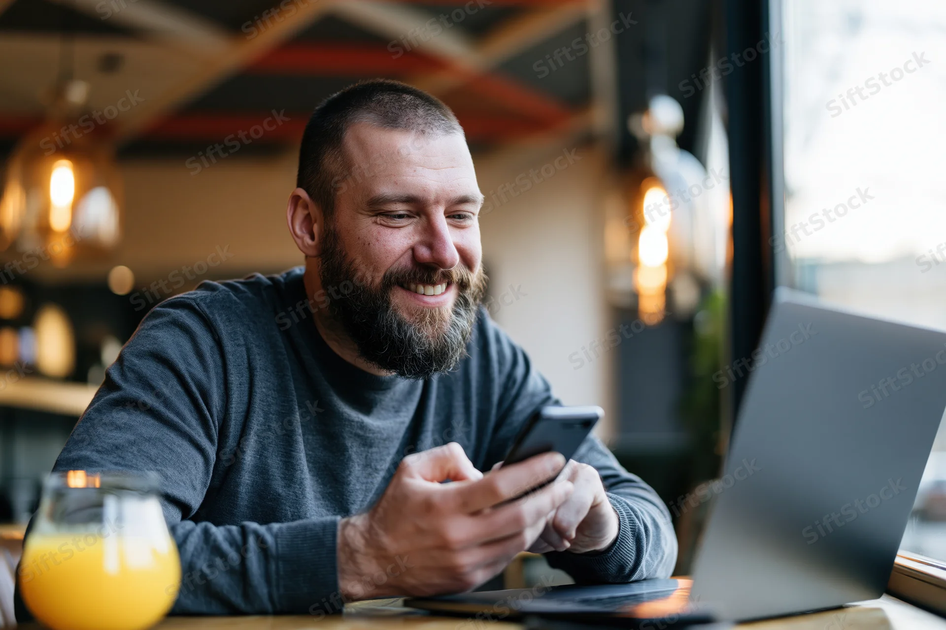 Bearded man smiling while using smartphone at cafe table with laptop and orange juice