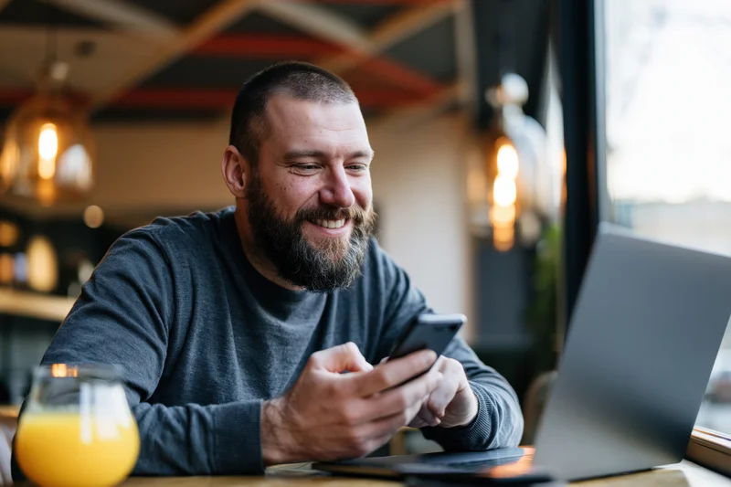 Bearded man smiling while using smartphone at cafe table with laptop and orange juice