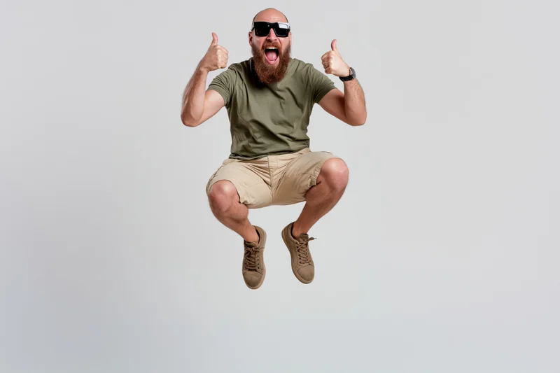 Bearded man mid-jump giving two thumbs up with mouth open in excitement against white background