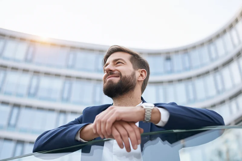 Bearded young businessman leaning on glass railing outside modern office building smiling upward