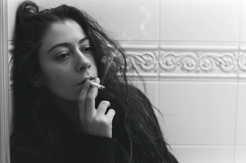 Young woman smoking a cigarette in a tiled bathroom, leaning against the wall with a contemplative expression