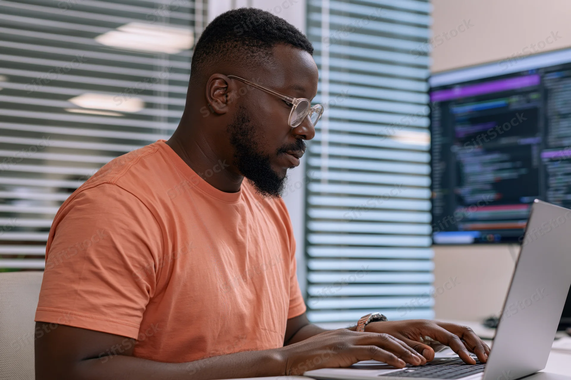 Black man typing on laptop at office desk with monitor displaying code
