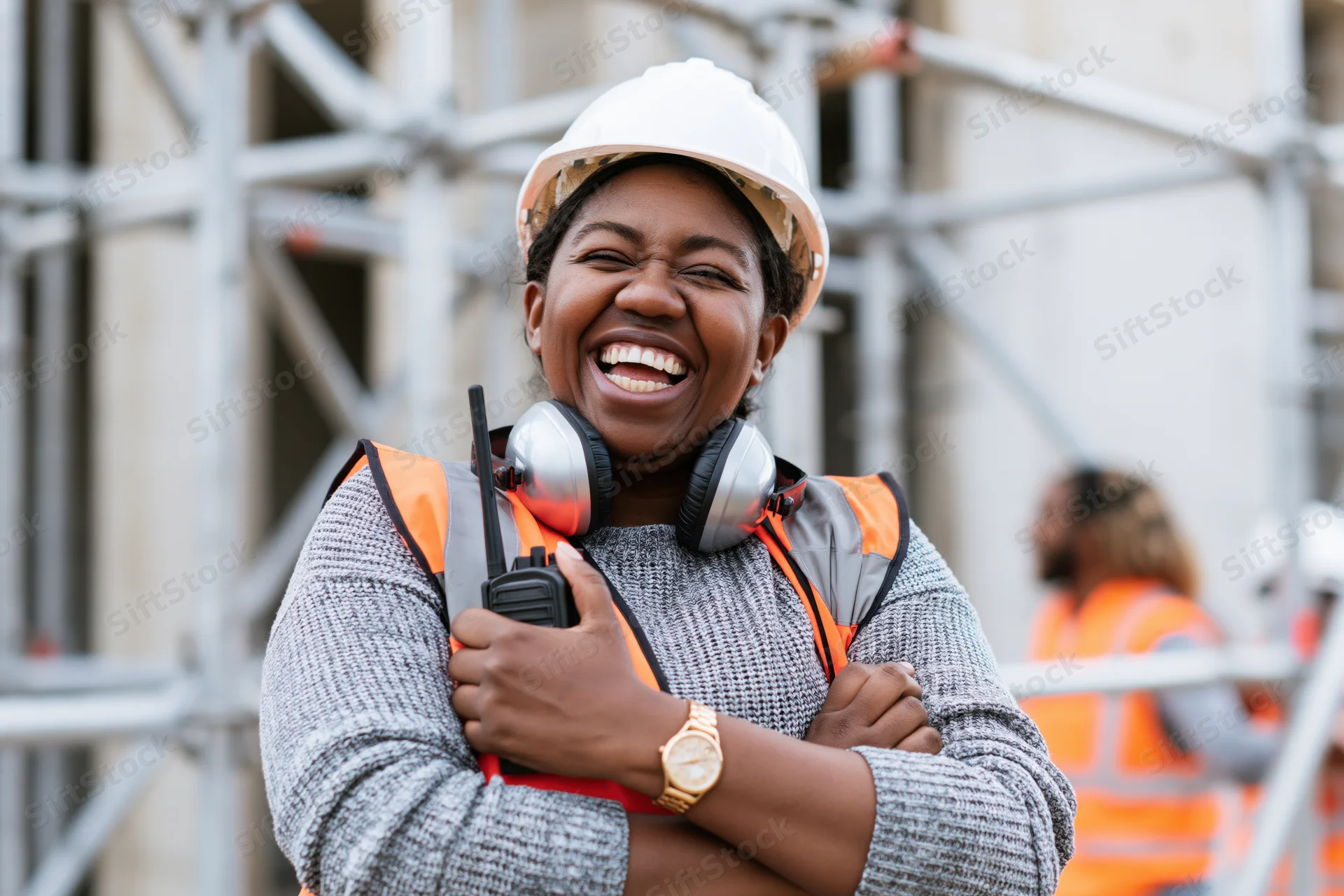 Black woman construction worker laughing with arms crossed on a building site wearing safety helmet and vest