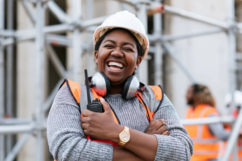 Black woman construction worker laughing with arms crossed on a building site wearing safety helmet and vest
