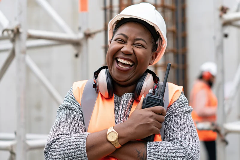 Black woman construction worker laughing while holding walkietalkie on building site
