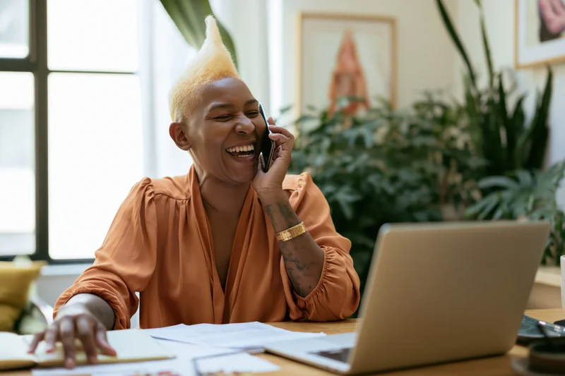 Black woman laughing while talking on phone at office desk with laptop and papers