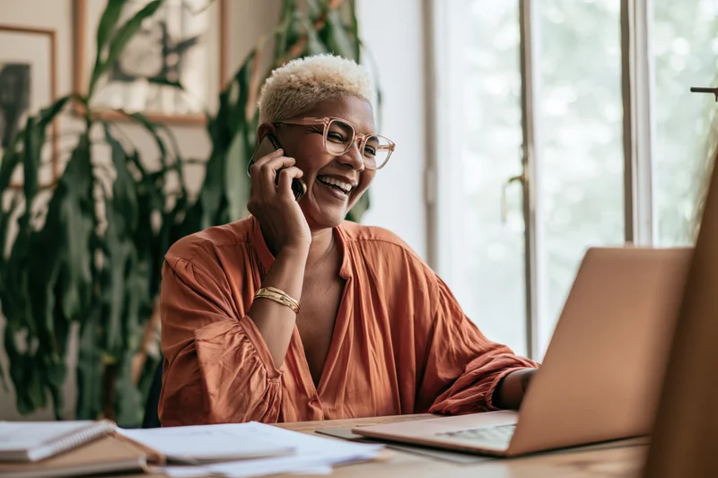 Black woman laughing while talking on phone at desk with laptop and plants nearby