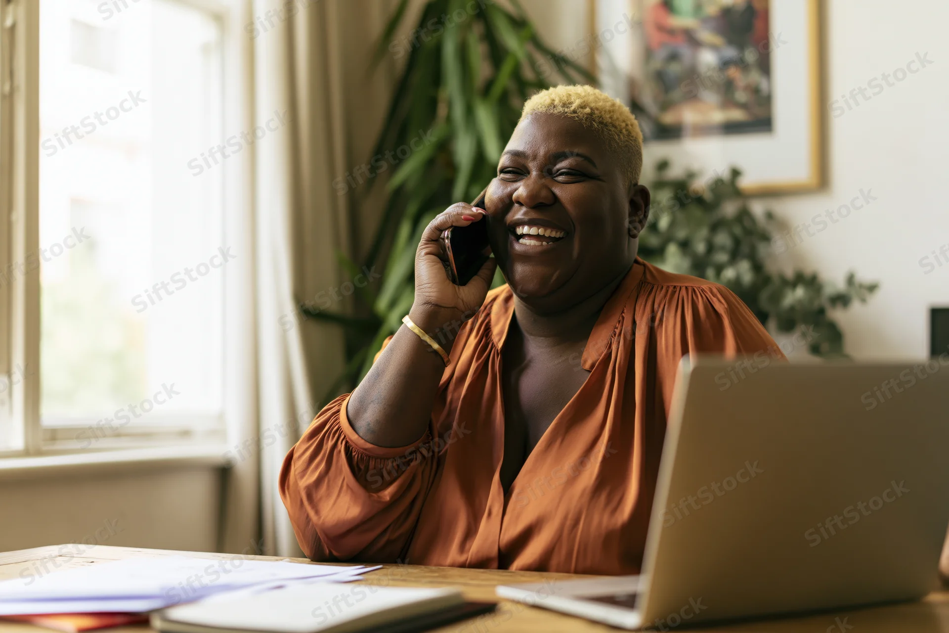 Black woman laughing while talking on phone at desk with laptop and papers near window