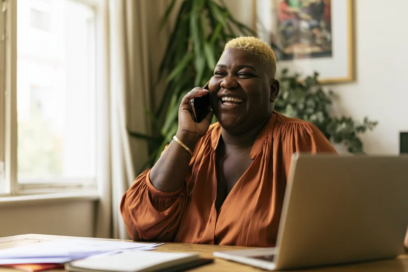 Black woman laughing while talking on phone at desk with laptop and papers near window