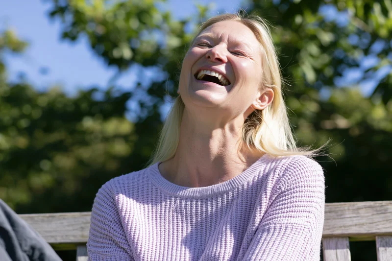 Blonde woman laughing openly with head tilted back on park bench in sunlight