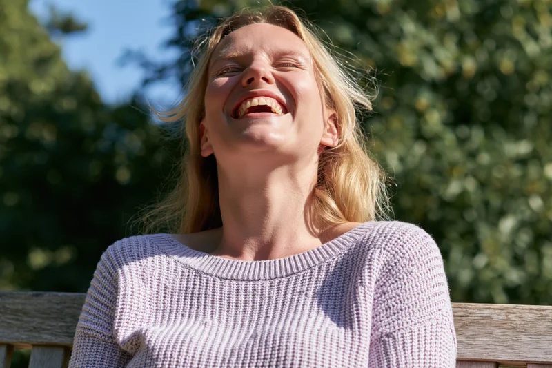 Blonde woman laughing openly with head tilted back on park bench in sunlight