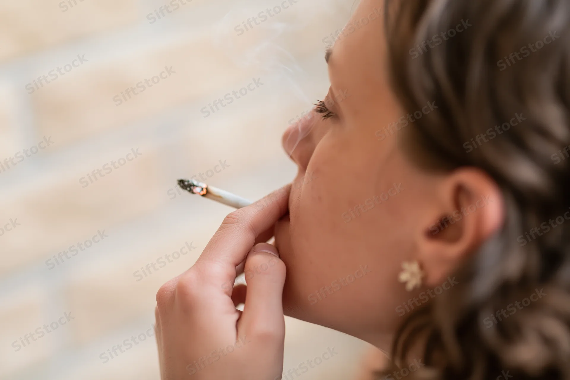 Young woman in profile smoking a cigarette, hand holding lit tobacco near her lips, soft indoor background