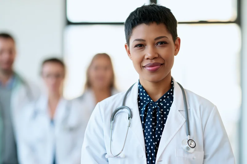 Young black female doctor smiling in lab coat with stethoscope, standing in front of medical team in clinic