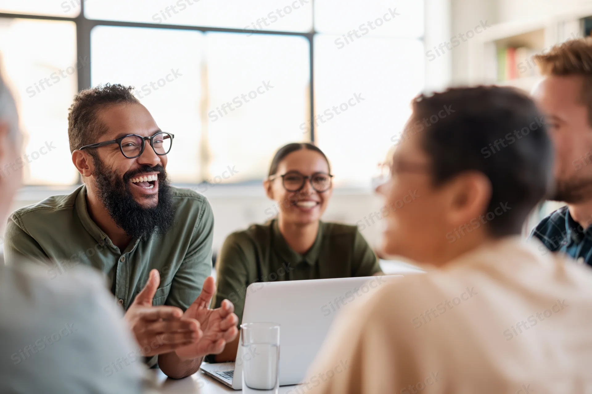 Diverse team of colleagues laughing and discussing during a casual office meeting around a table with a laptop and glass of water