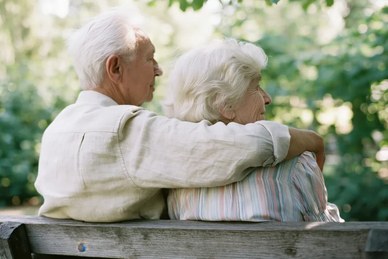 Elderly couple sitting close together on a weathered wooden bench in a park, man’s arm around woman’s shoulder, both facing right, surrounded by green foliage and sunlight