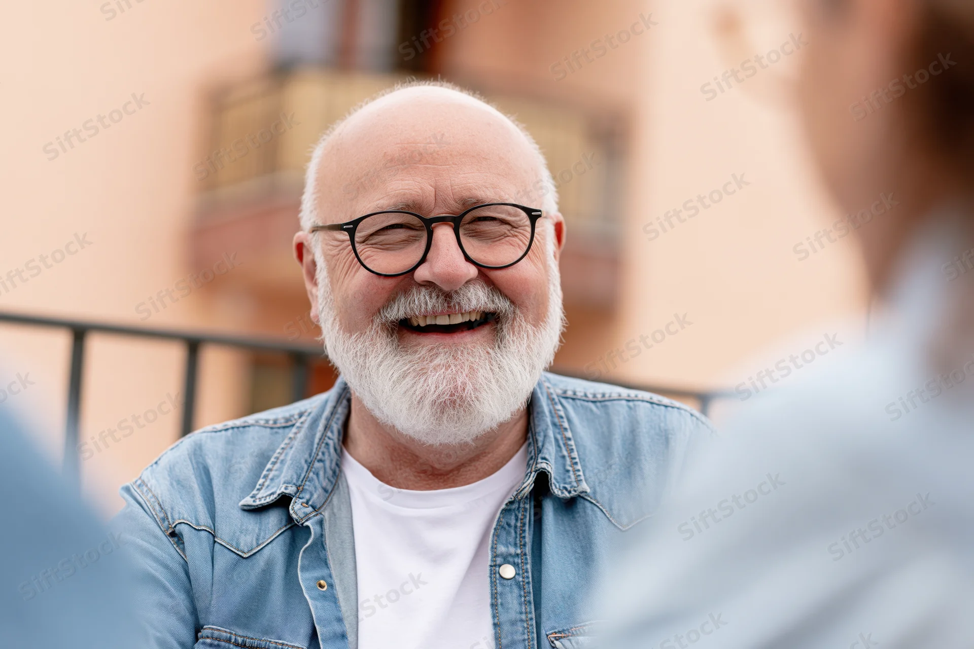 Senior man with white beard and glasses laughing warmly outdoors on a balcony