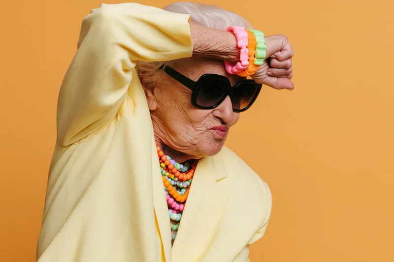 Elderly woman in yellow blazer doing dab pose with sunglasses and colorful jewelry against orange background