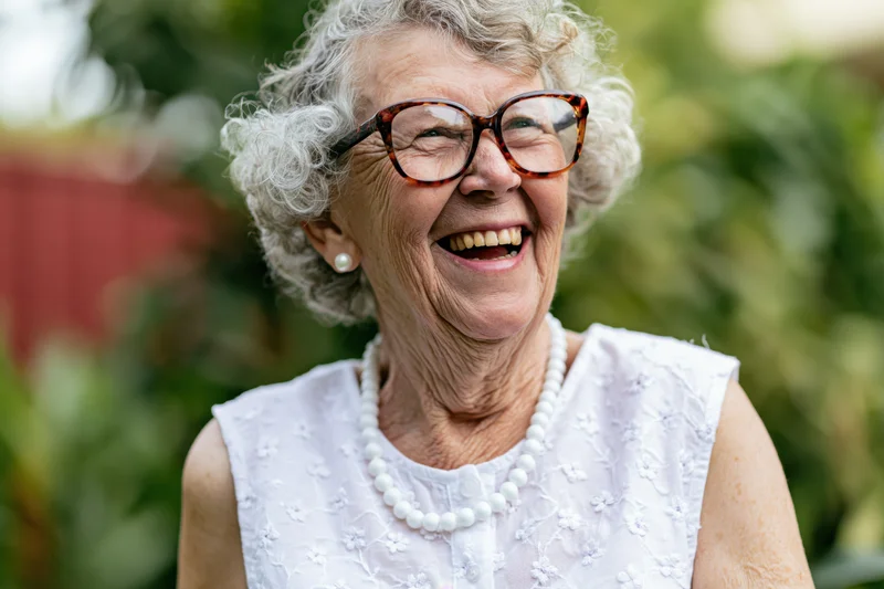 Elderly woman laughing with tortoiseshell glasses and pearl necklace in a garden setting