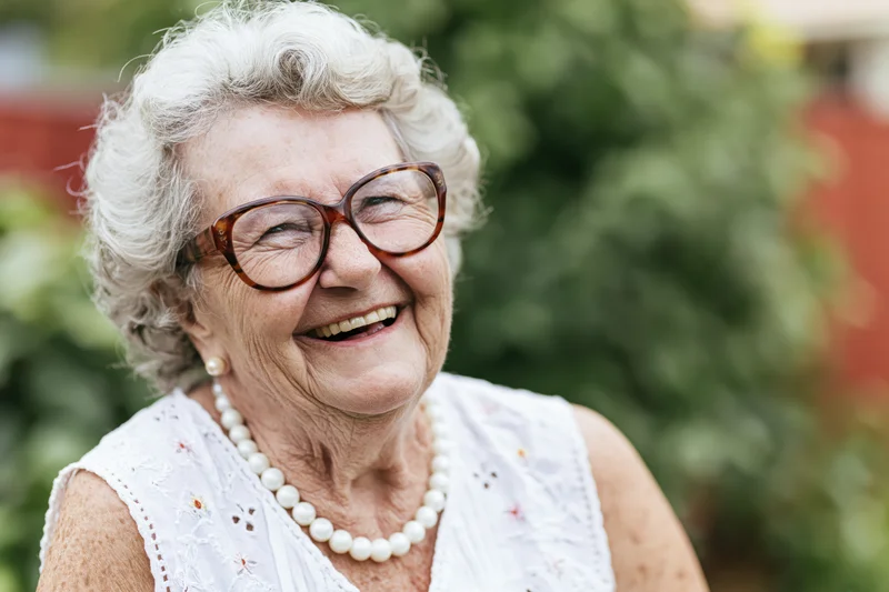 Elderly woman smiling warmly outdoors wearing glasses and pearl necklace