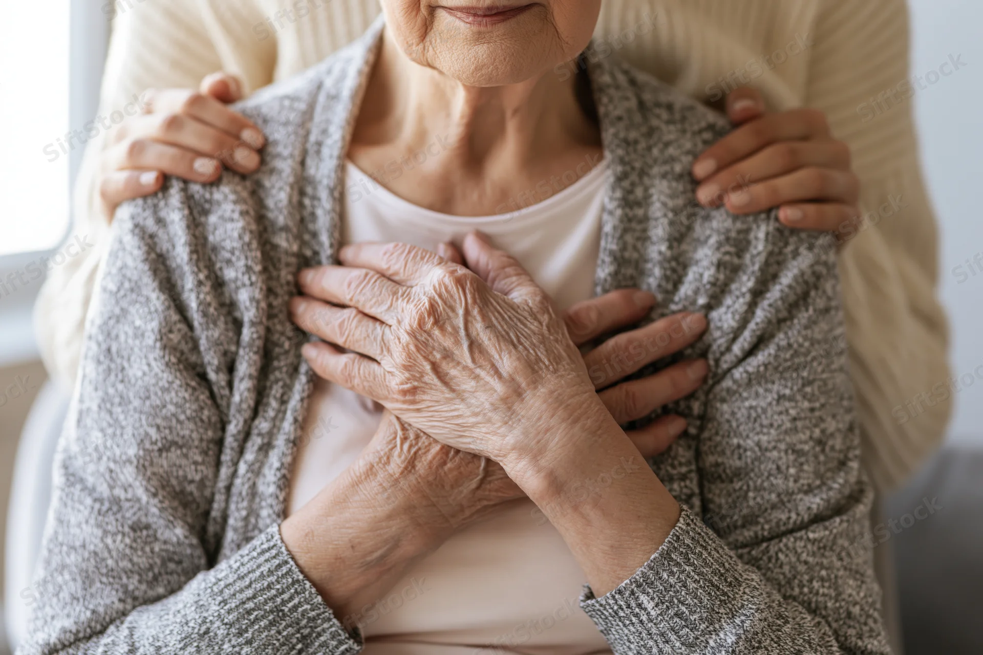 Elderly woman with hands clasped over chest, receiving supportive touch from another person seated behind her