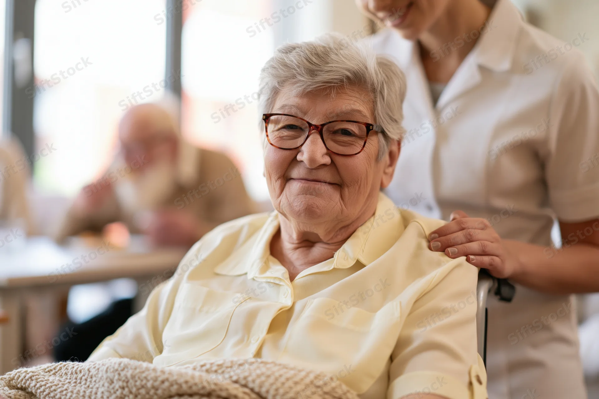Elderly woman smiling in wheelchair with caregiver’s hand on her shoulder in a bright indoor setting