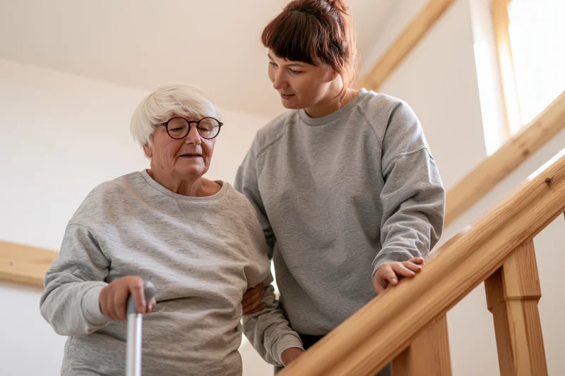 Elderly woman with short white hair and round glasses descending stairs with caregiver support, holding cane and handrail in home setting