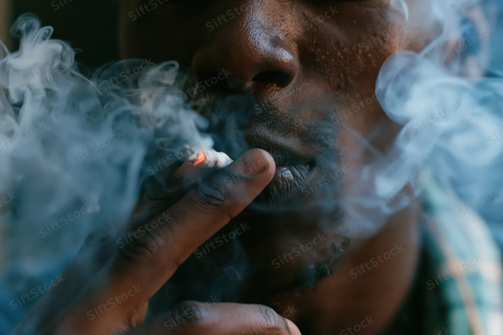 Young black man smoking a cigarette, close-up on lower face and hand, smoke swirling around his mouth